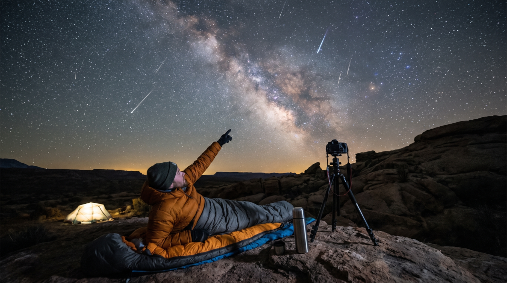 Persona en saco de dormir bajo cielo estrellado con Vía Láctea y meteoros. Tienda de campaña iluminada, cámara en trípode, termo y taza.