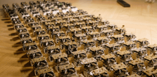 Pequeños robots con componentes electrónicos expuestos alineados en filas diagonales sobre un suelo de madera.