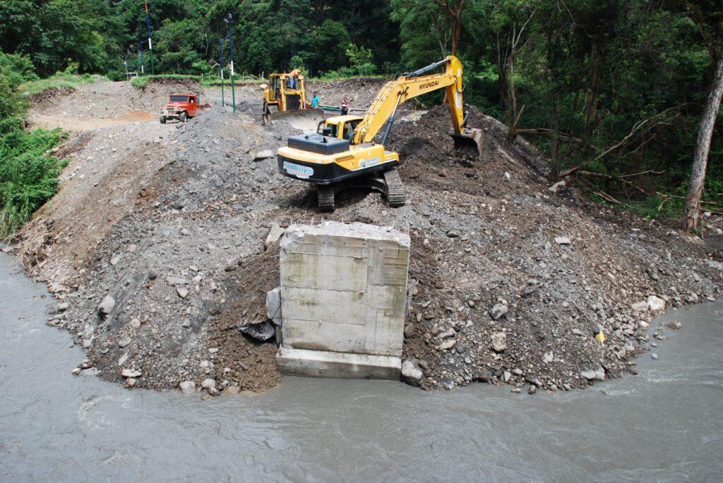 Vista elevada del estribo del puente de Ospino expuesto en el cauce y de una excavadora sobre el relleno de tierra.